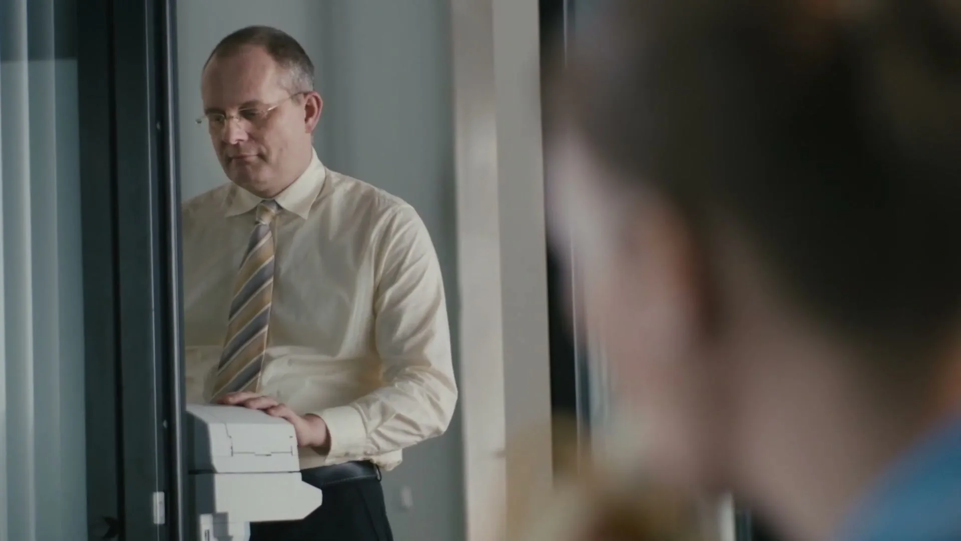A middle-aged man in a shirt and tie stands by a printer, looking at a woman whose back is to the camera in an office.