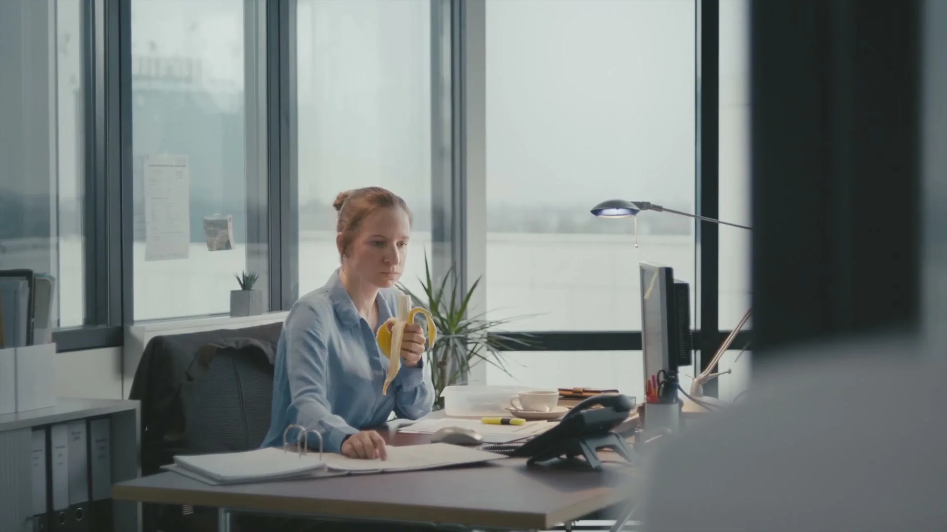 A young woman in a blue shirt sits at an office desk, eating a banana and looking directly at the camera in a modern office with large windows.