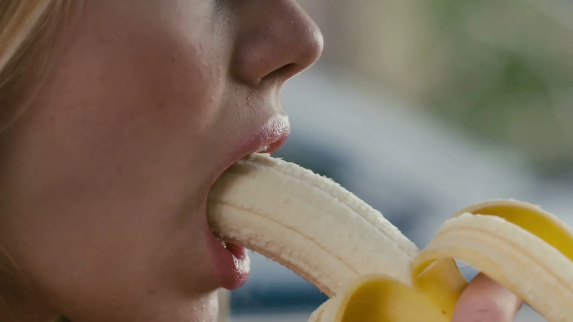 A young woman with blonde hair and pink lipstick is seen in a closeup, suggestively eating a banana.
