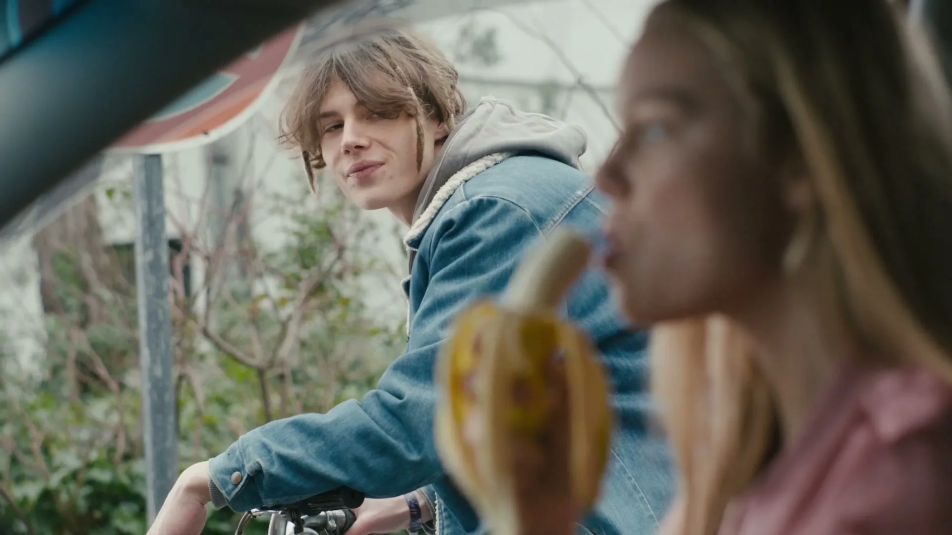 A young woman in a car eats a banana while a young man on a bicycle looks at her from outside the car on a street.