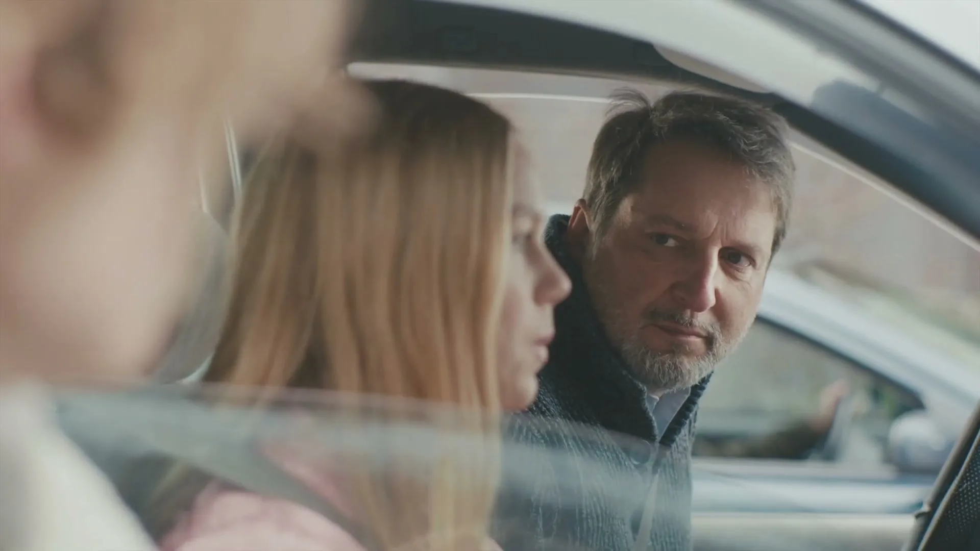 A man with a beard and a woman with long blonde hair are sitting in a car, with the man looking at the camera and the woman looking away.