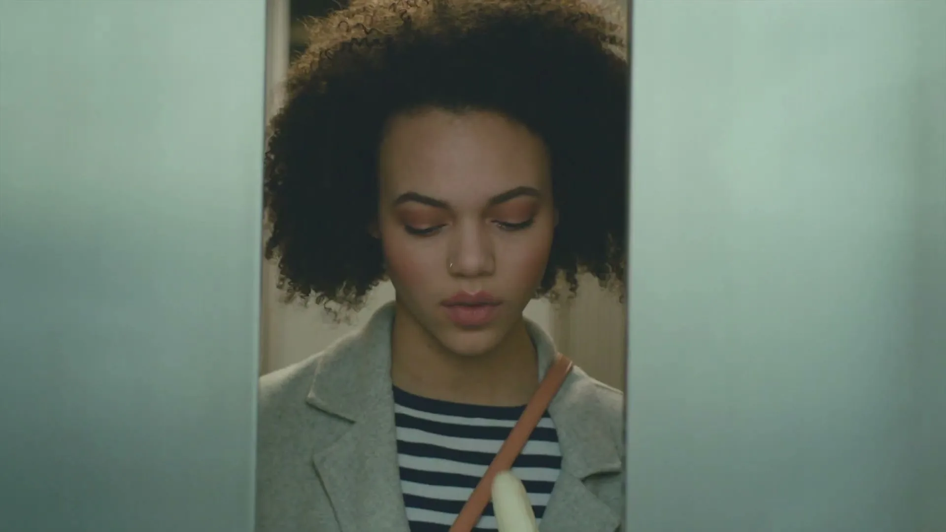 A young woman with curly hair, wearing a striped shirt and a grey coat, is seen through a closing door, then eats a banana in an elevator.