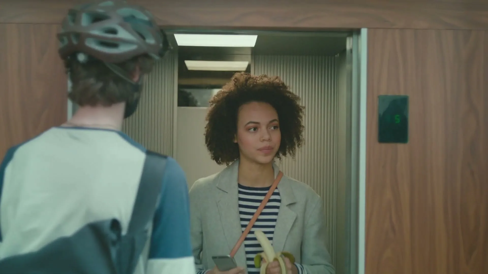 A young woman with curly hair, wearing a striped shirt and blazer, eats a banana while looking at a man in an elevator.