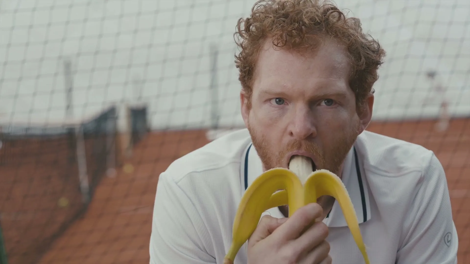 A man with red hair and a beard, wearing a white polo shirt, eats a banana suggestively on a tennis court.