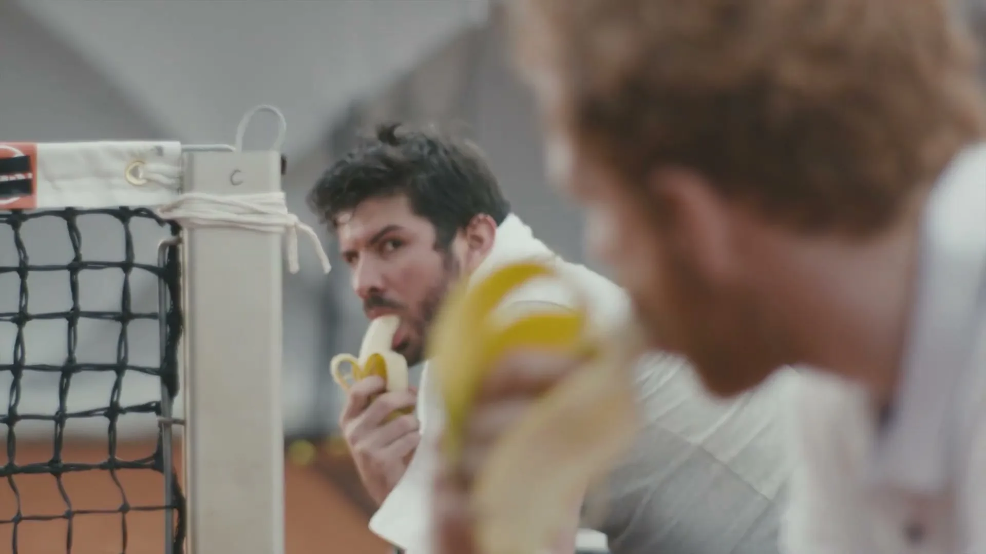 Two male tennis players, one with red hair and a beard, eat bananas on a tennis court, with the red-haired man in the foreground.