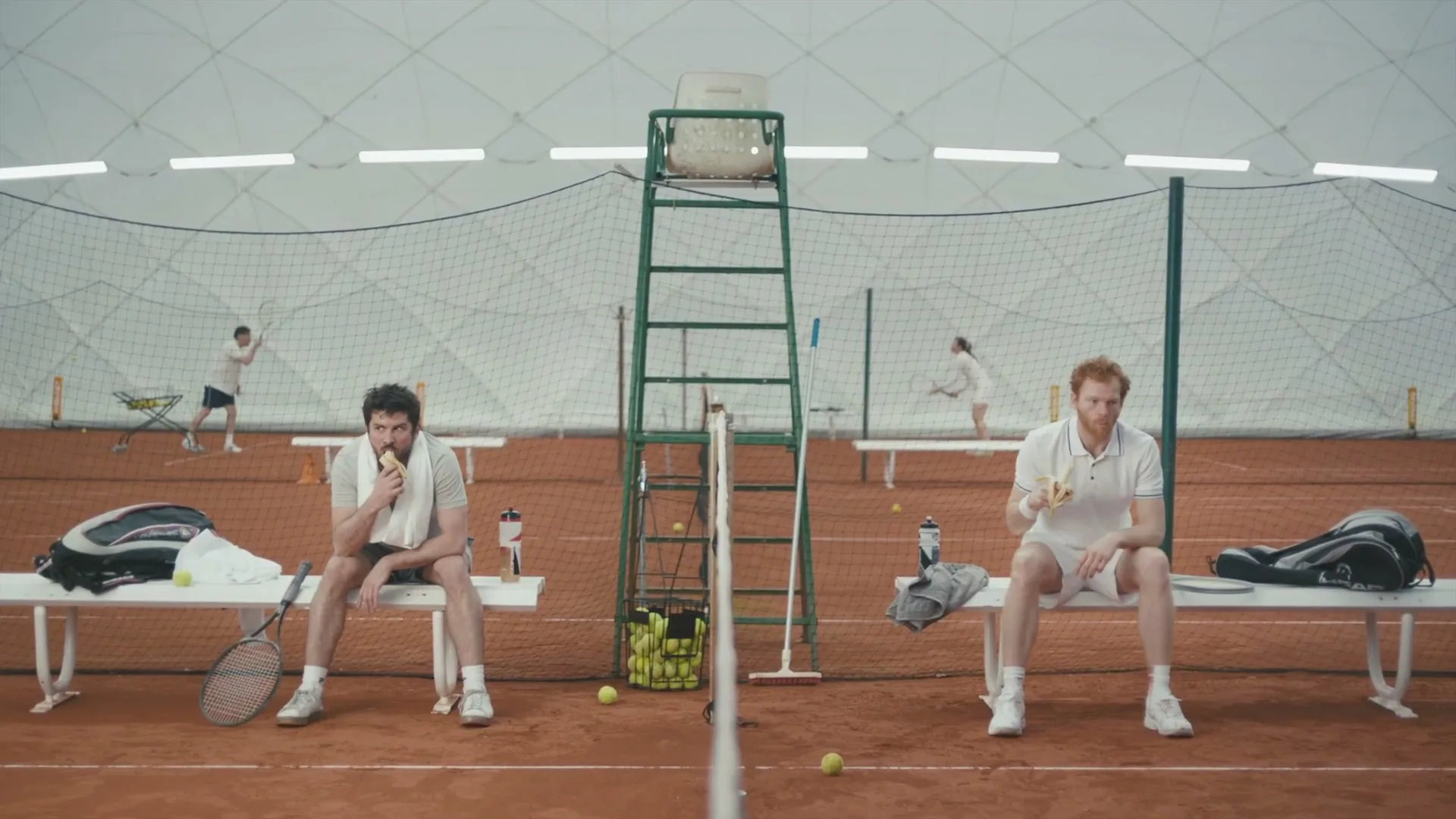 Two male tennis players sit on benches on a clay court, eating bananas, with other players in the background.