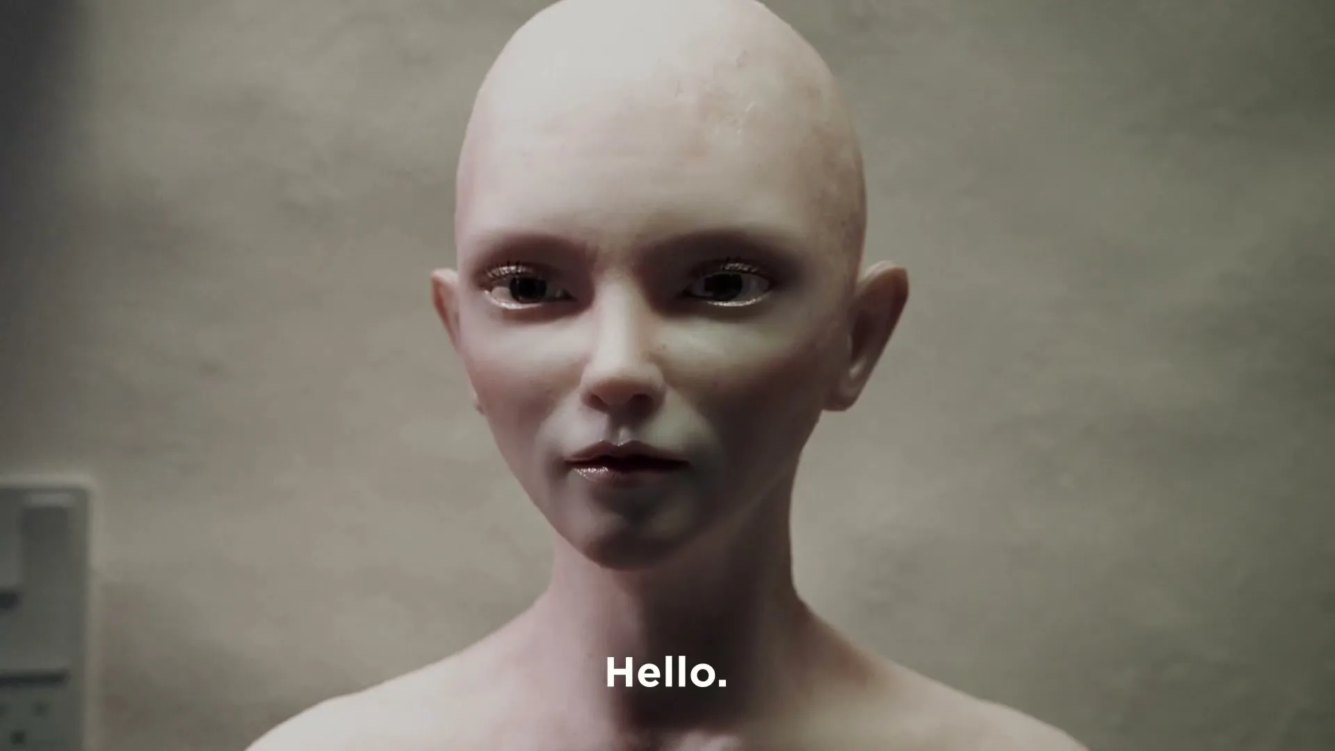 A bald, pale, androgynous figure with large eyes and red lips slowly opens their eyes and looks around in a dimly lit room.
