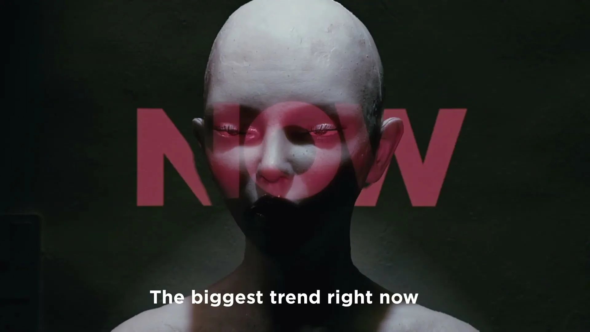 A bald, pale, androgynous mannequin-like figure with red light projected onto its face, stands in a dark room as text appears and disappears.