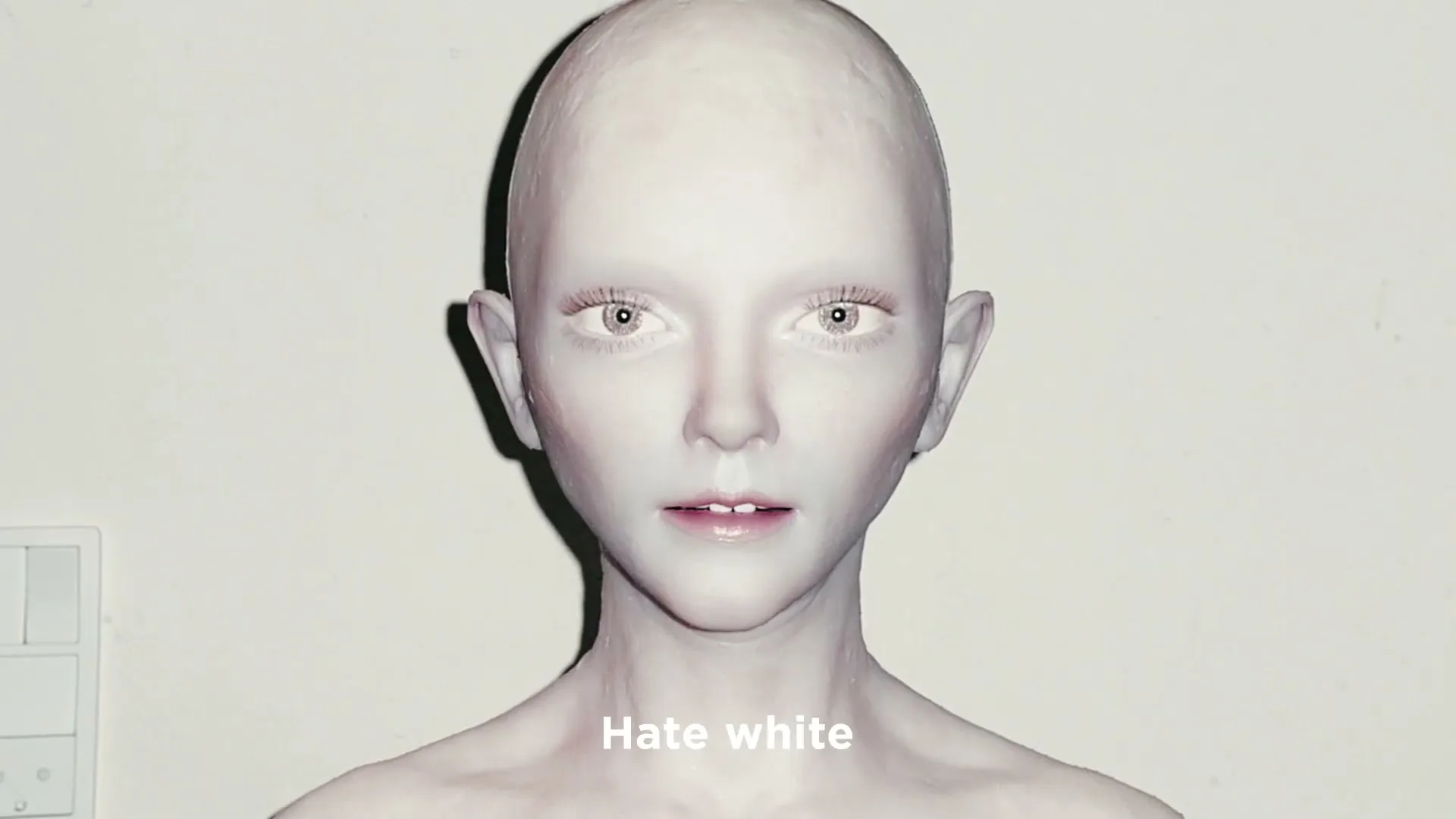 A pale, bald, androgynous figure with glowing eyes emerges from darkness, looking directly at the camera with a neutral expression.