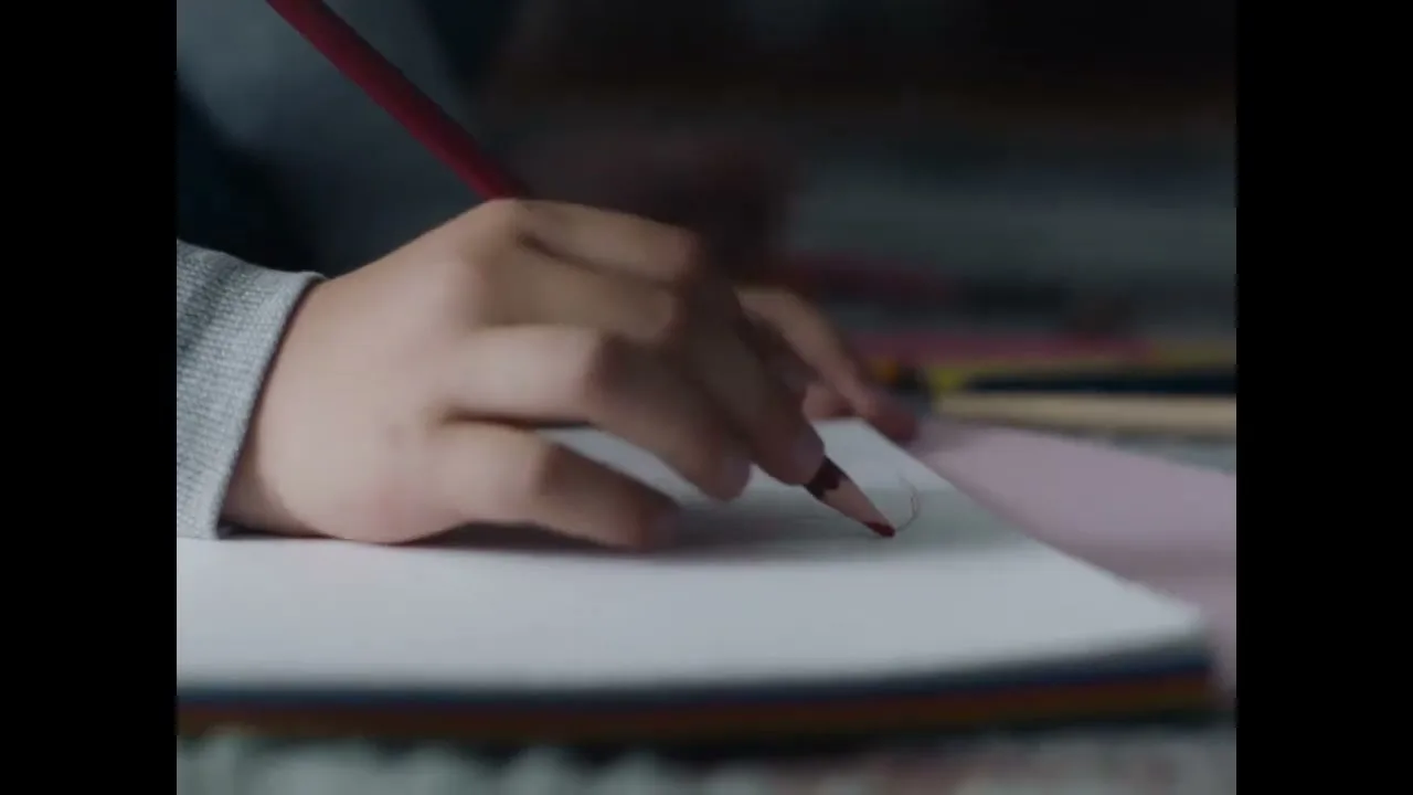 A child's hand in a grey sleeve draws a red circle on white paper with a red pencil, in a close-up shot.