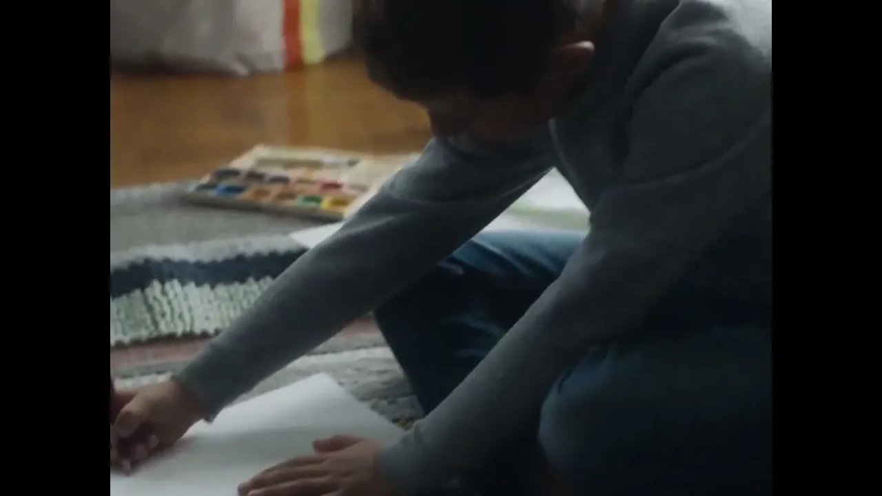 A young boy sits on a rug, drawing on paper with colored pencils, with a watercolor set nearby.