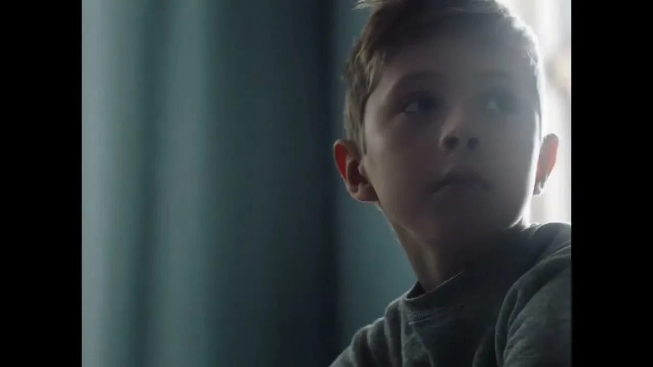 A young boy with light brown hair, wearing a grey sweatshirt, looks up and to the right in a dimly lit room.