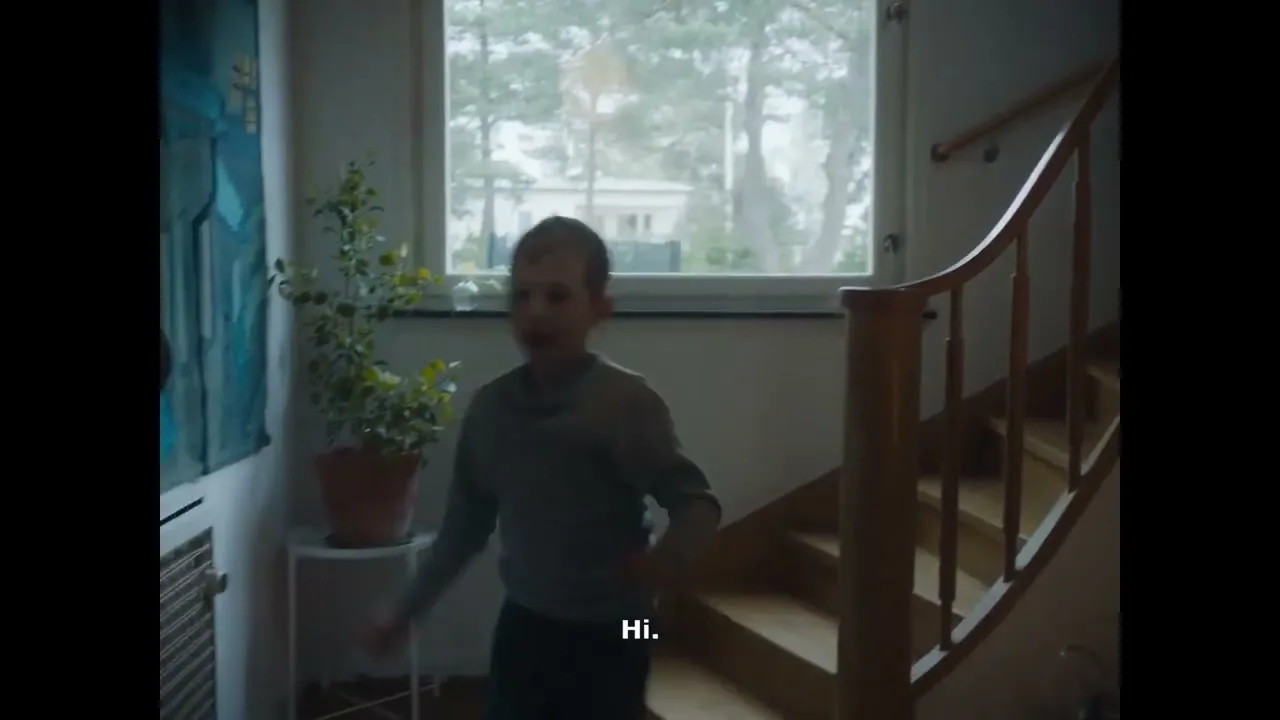A young boy in a grey shirt and blue pants runs down a wooden staircase in a brightly lit home, looking excited.