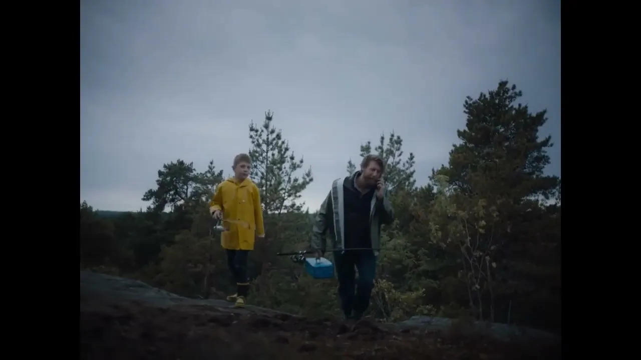 A man and a boy walk through a forest with fishing gear, the man talking on a phone, under a cloudy sky.