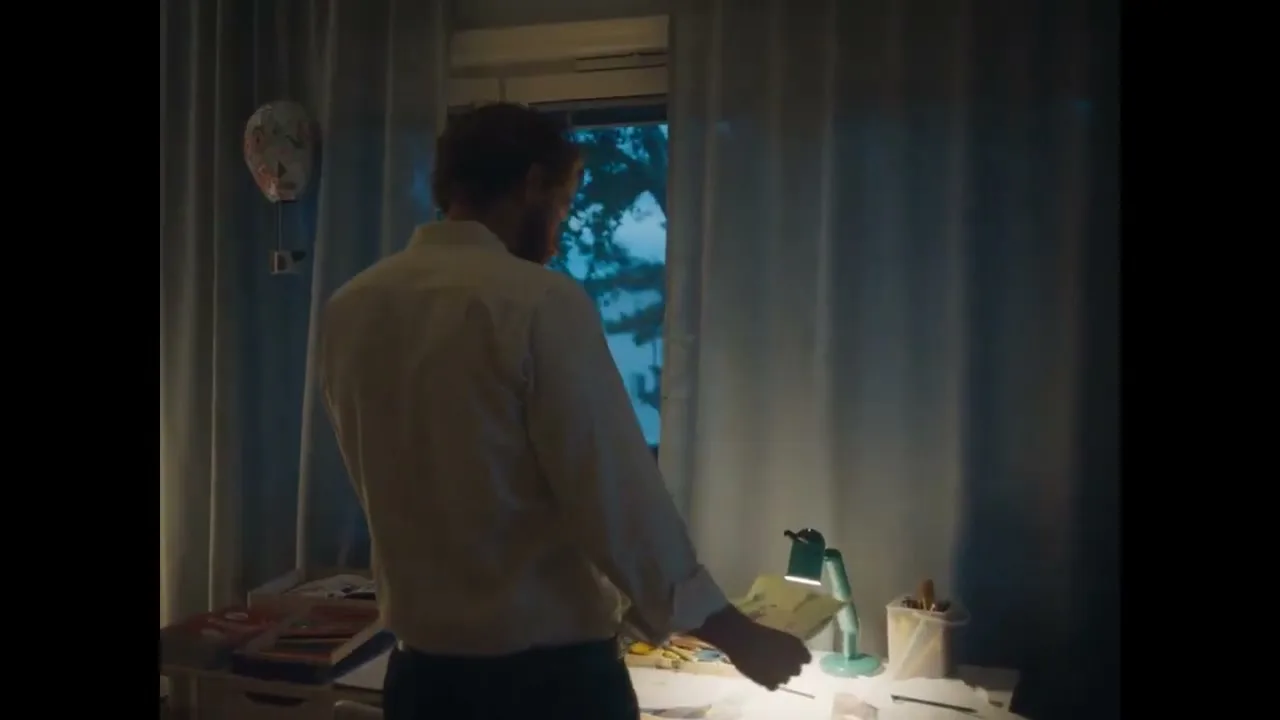 A man with a beard and white shirt stands at a desk, looking out a window at dusk in a bedroom.