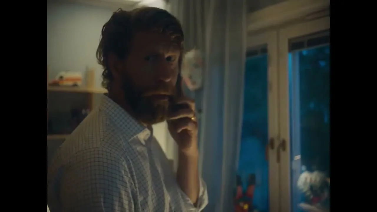 A man with a beard talks on the phone, looking concerned, in a dimly lit room at night.