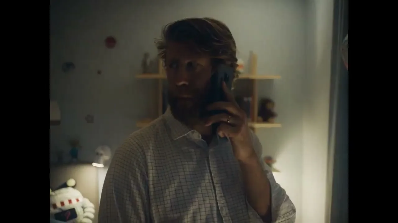 A man with a beard and a plaid shirt talks on a phone in a dimly lit child's bedroom, looking around with a concerned expression.