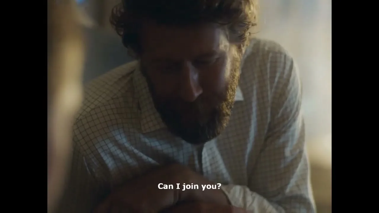 A man with a beard and a checkered shirt looks up with a hopeful expression, asking to join someone in a dimly lit interior.
