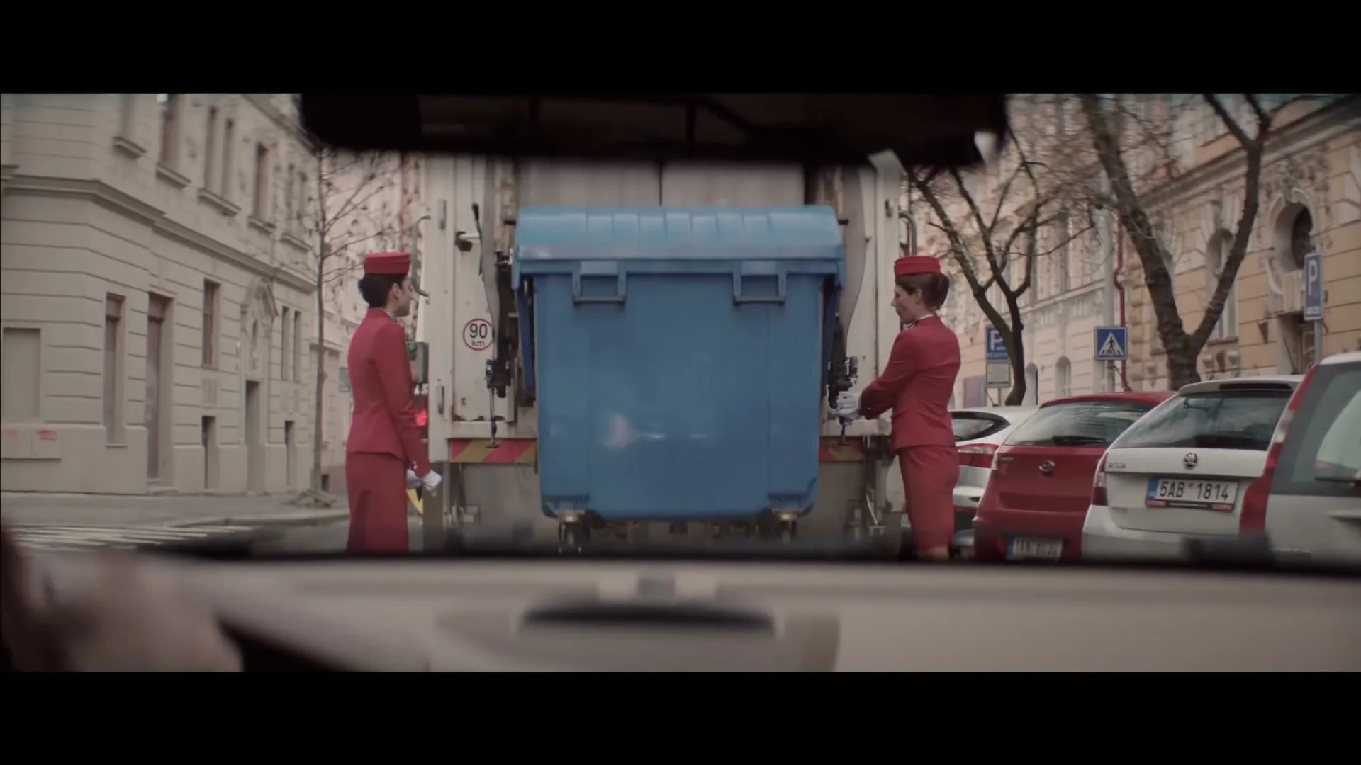 Two women in red flight attendant uniforms stand on either side of a blue dumpster being lifted by a garbage truck on a city street.