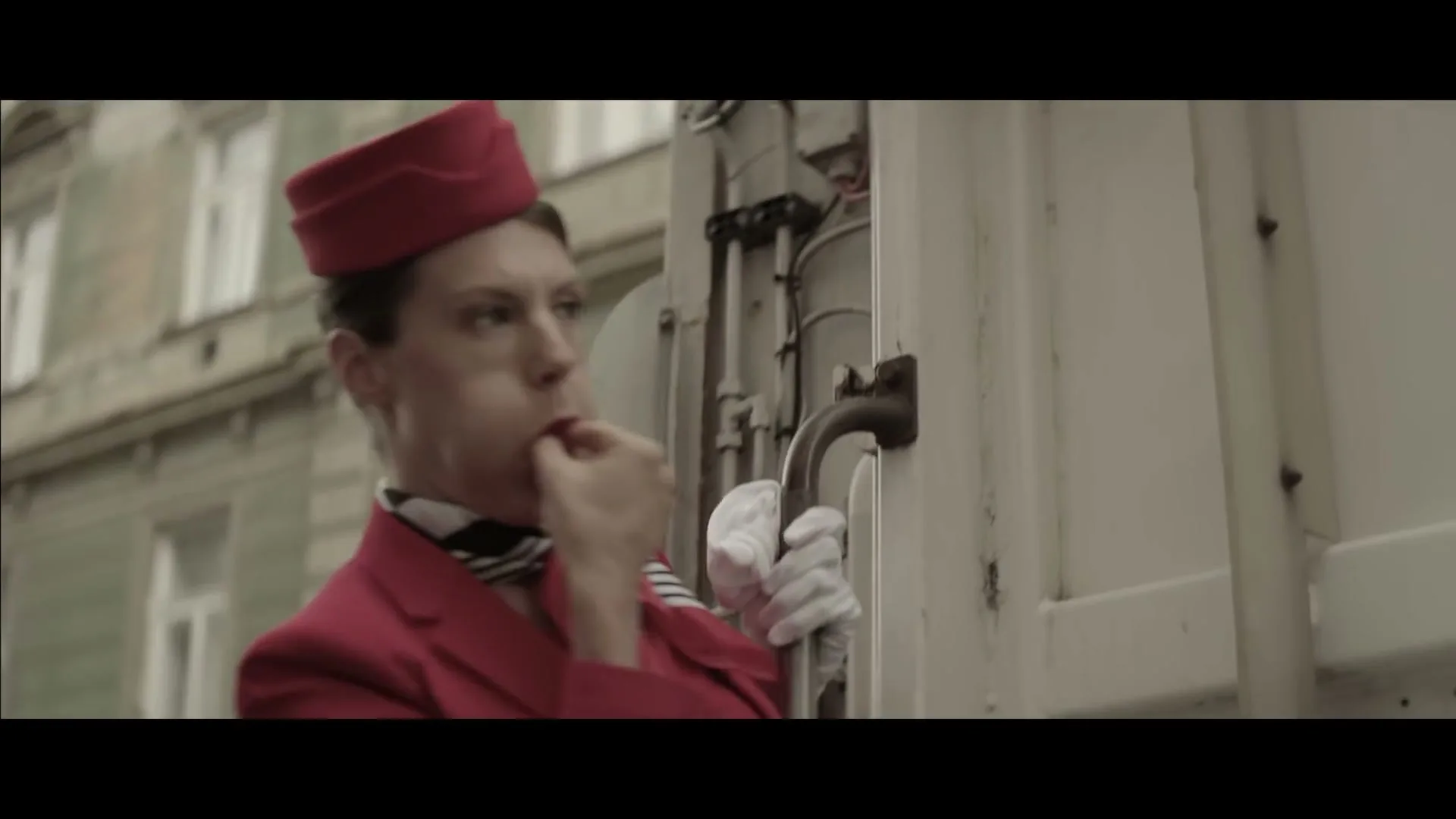 A female flight attendant in a red uniform and white gloves whistles and then spits out a piece of gum while holding onto the side of a garbage truck on a city street.