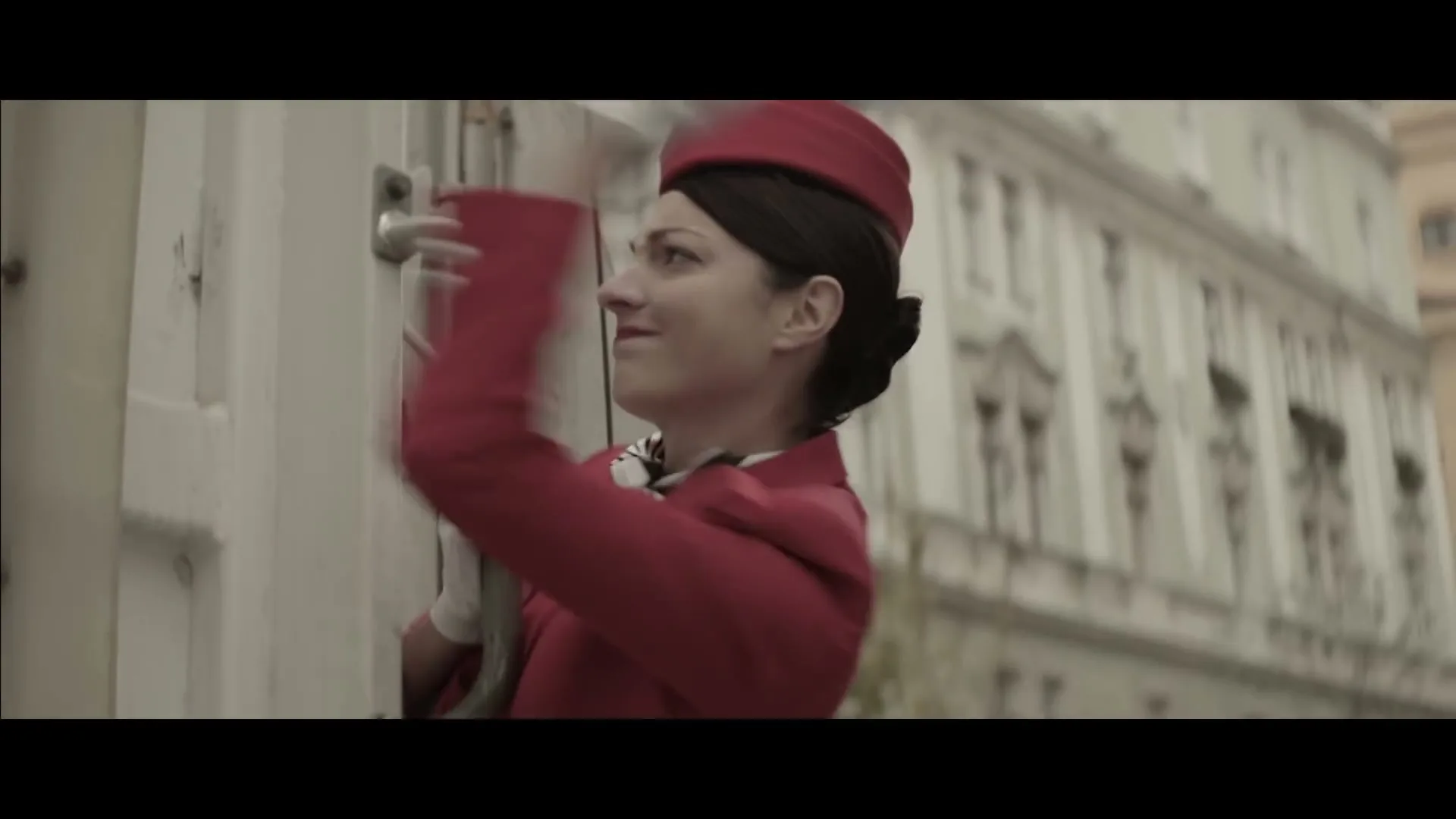 A female flight attendant in a red uniform and white gloves struggles to open a large, white metal door on a city street.