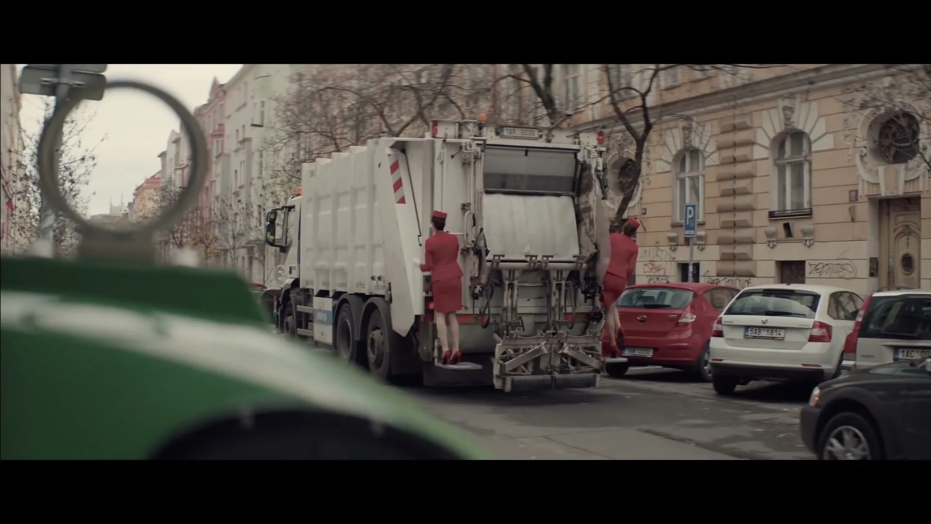 Two women in red flight attendant uniforms are riding on the back of a white garbage truck, which is driving down a city street lined with parked cars and buildings.