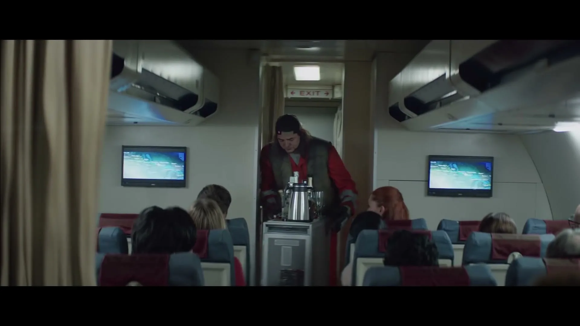 A male flight attendant, dressed in a red shirt and grey vest, pushes a beverage cart down the aisle of an airplane, serving passengers.