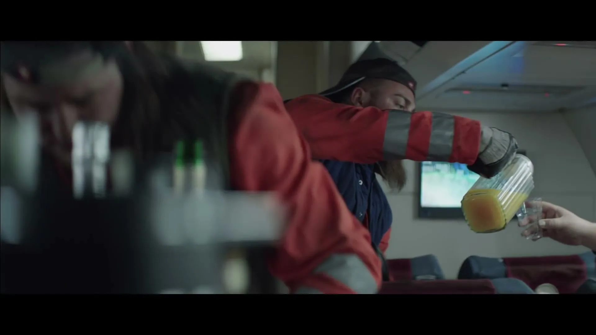 A male garbage collector, wearing a cap and work uniform, pours orange juice from a pitcher into a passenger's glass on an airplane.