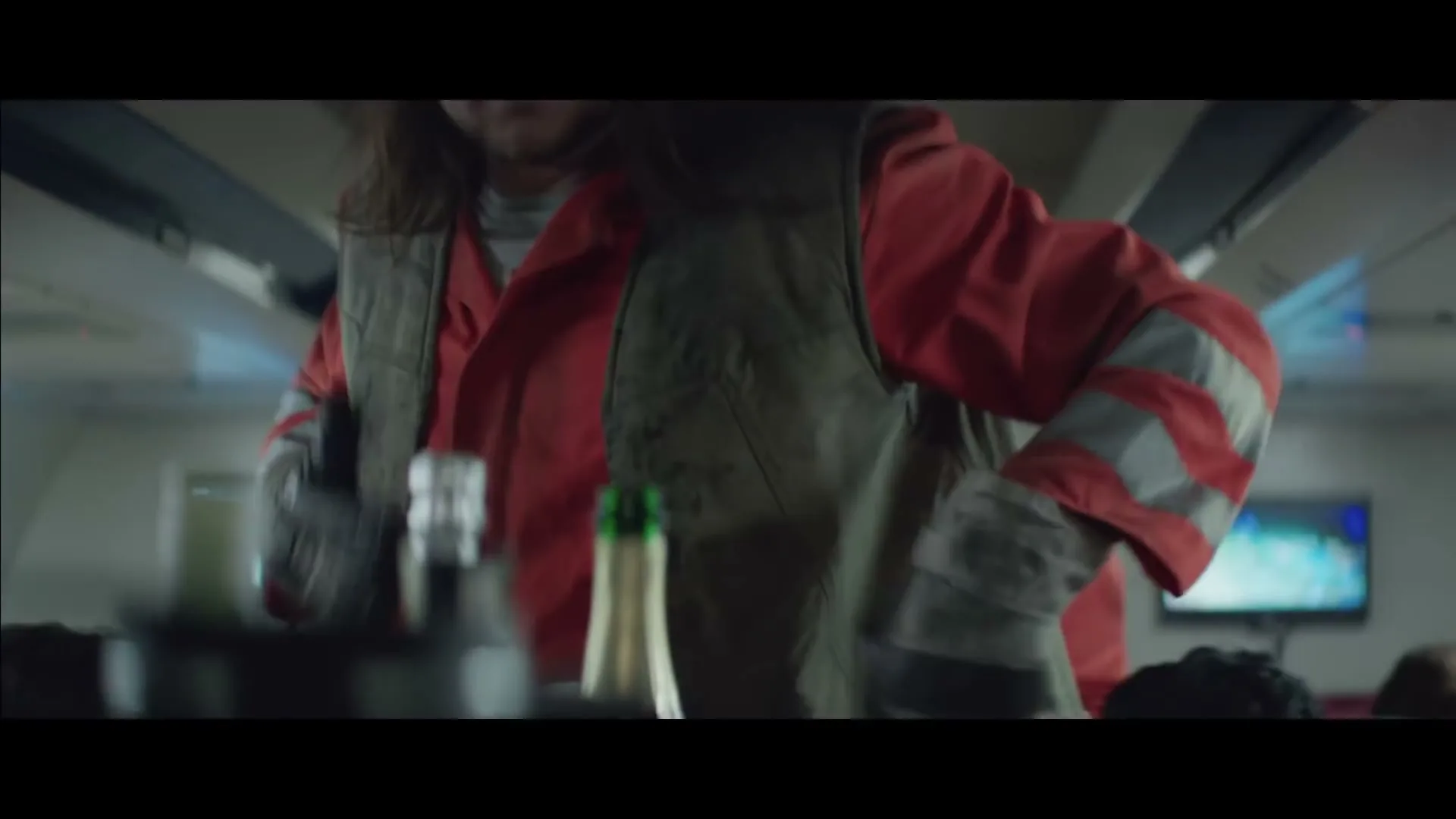 A female garbage collector in a red and grey uniform is seen from a low angle, bending over a cart with bottles on it, inside a plane.