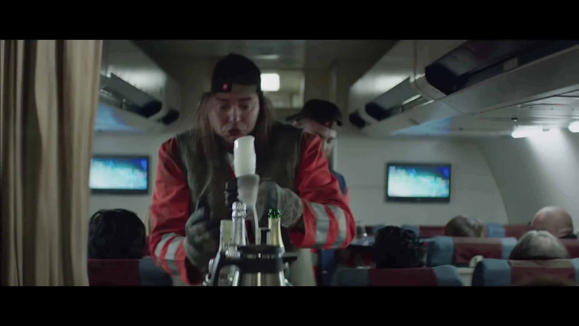 Two men dressed as garbage collectors serve drinks to passengers on an airplane, one pouring champagne and the other holding a glass of orange juice.