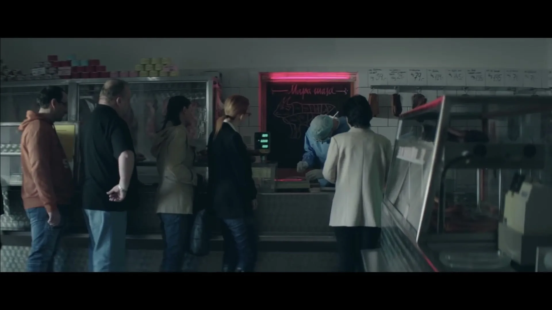 A group of people stand in line at a butcher shop, watching the butcher prepare meat behind the counter.