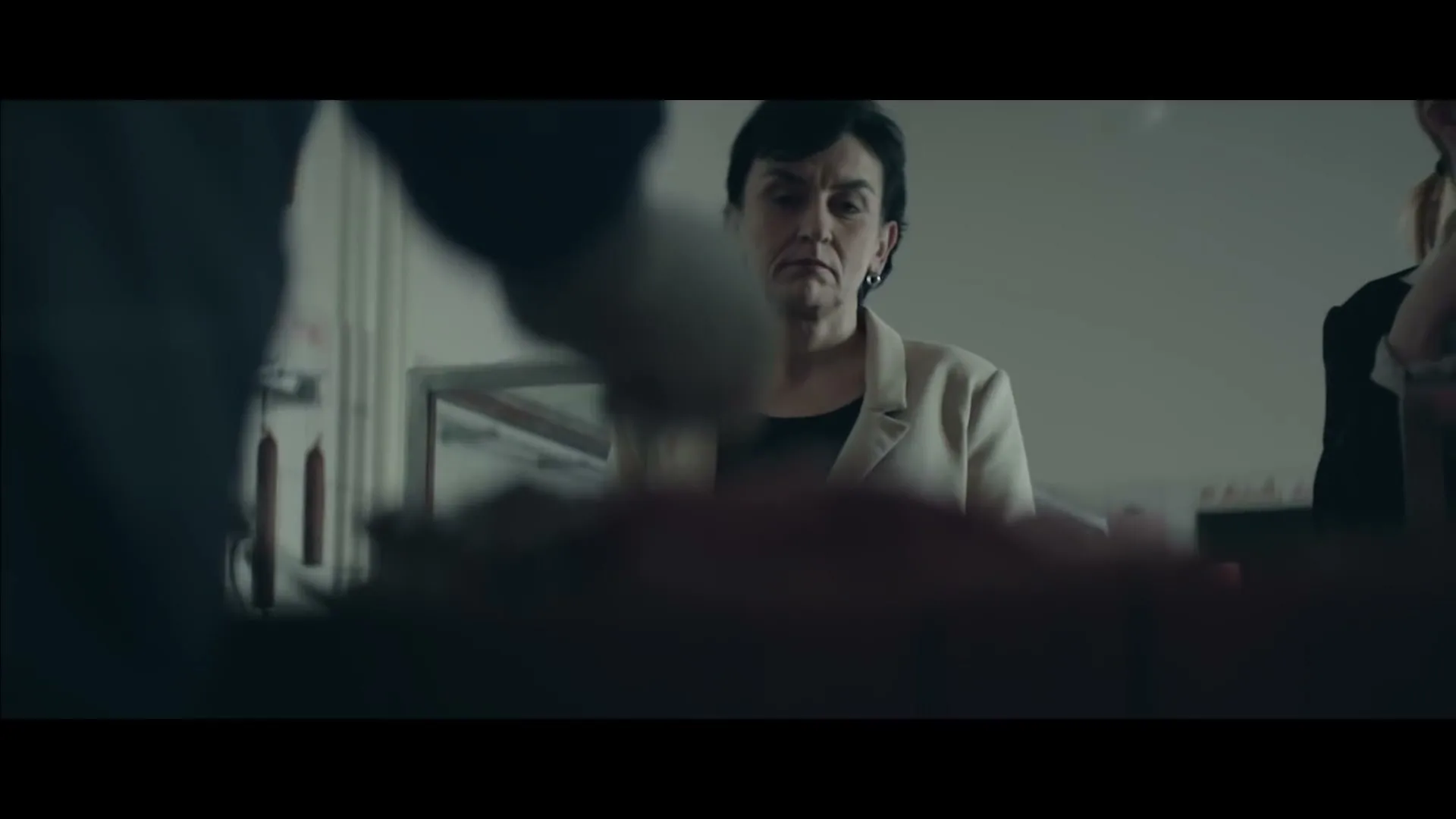 A middle-aged woman with dark hair in a beige blazer watches intently as a person in the foreground works on something out of focus in an interior setting.