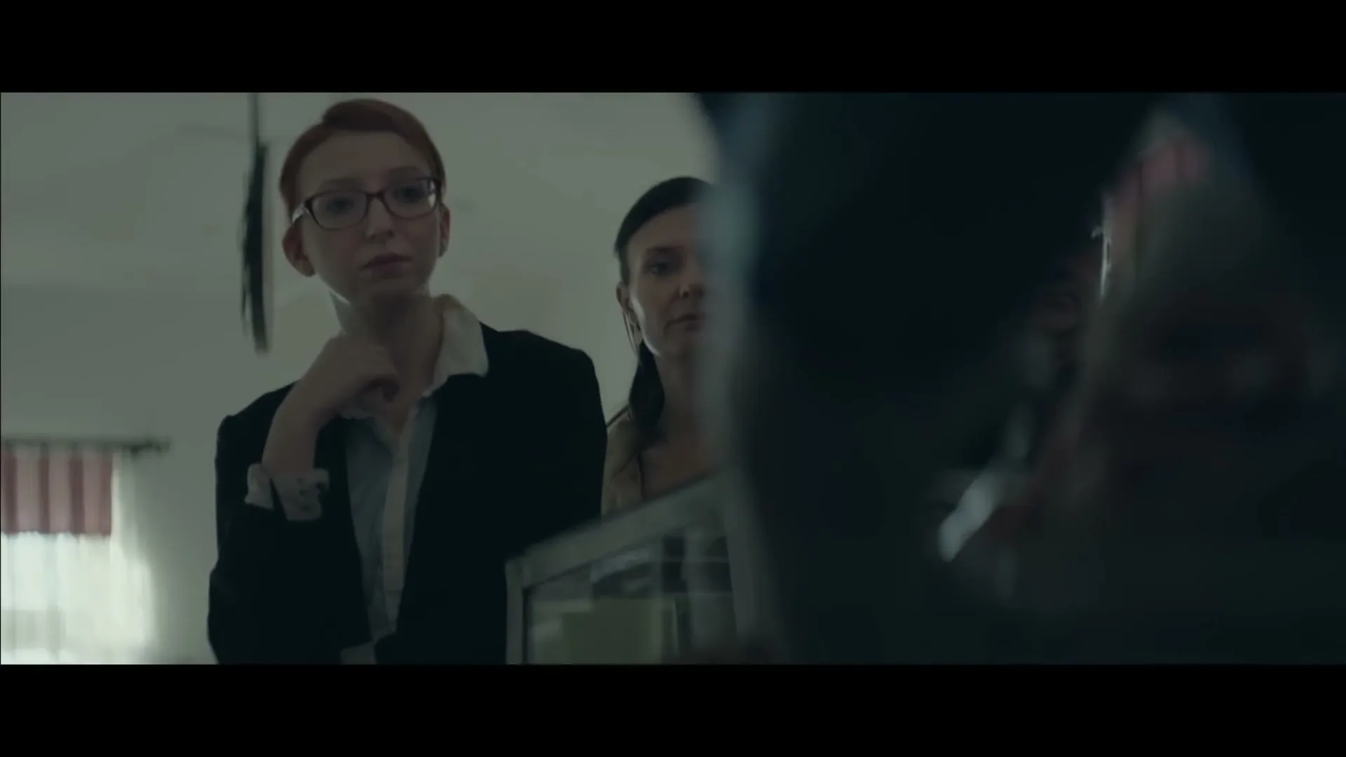Two women, one with red hair and glasses, the other with dark hair, stand behind a counter, looking intently at something off-screen in an interior setting.