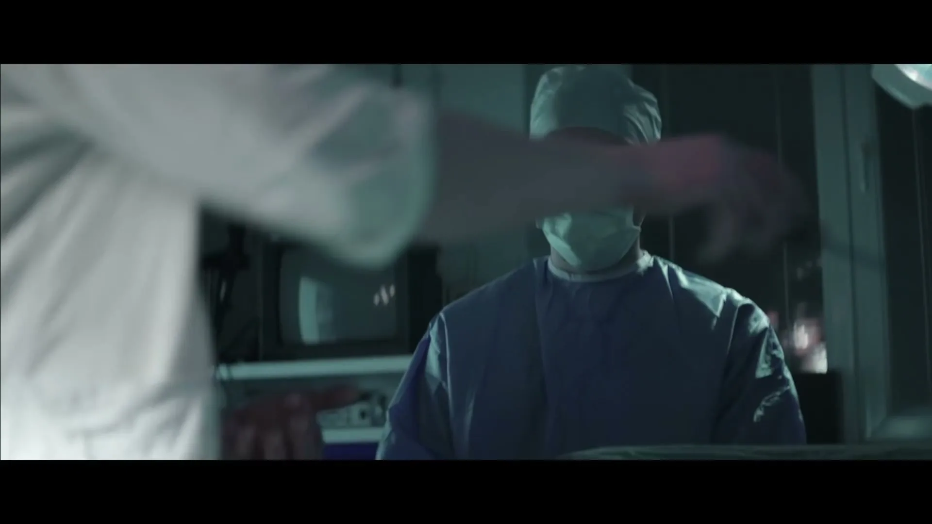 A surgeon in blue scrubs and a mask looks down while another person's arm reaches into the frame in a dimly lit operating room.