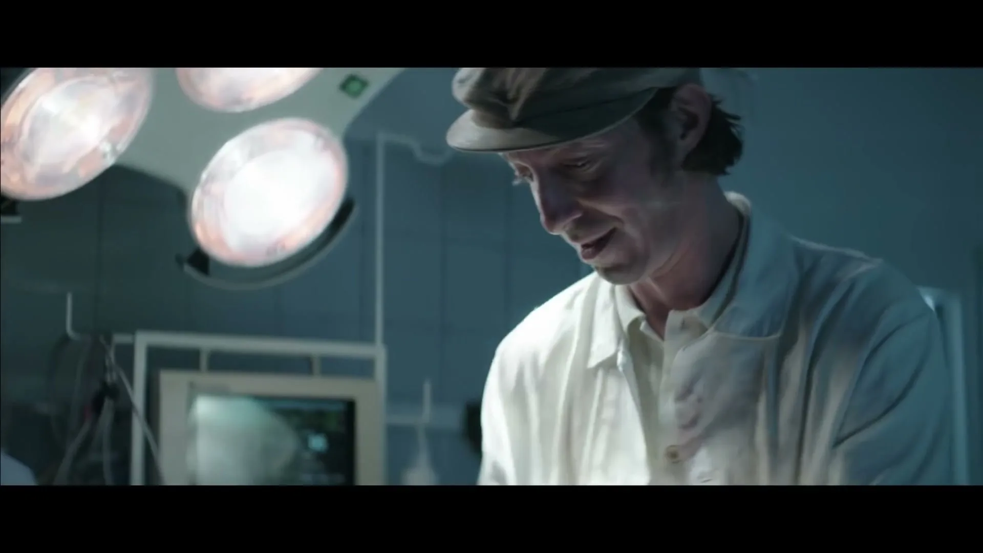 A man in a white coat and cap smiles while looking down, with surgical lights and medical equipment in the background of an operating room.