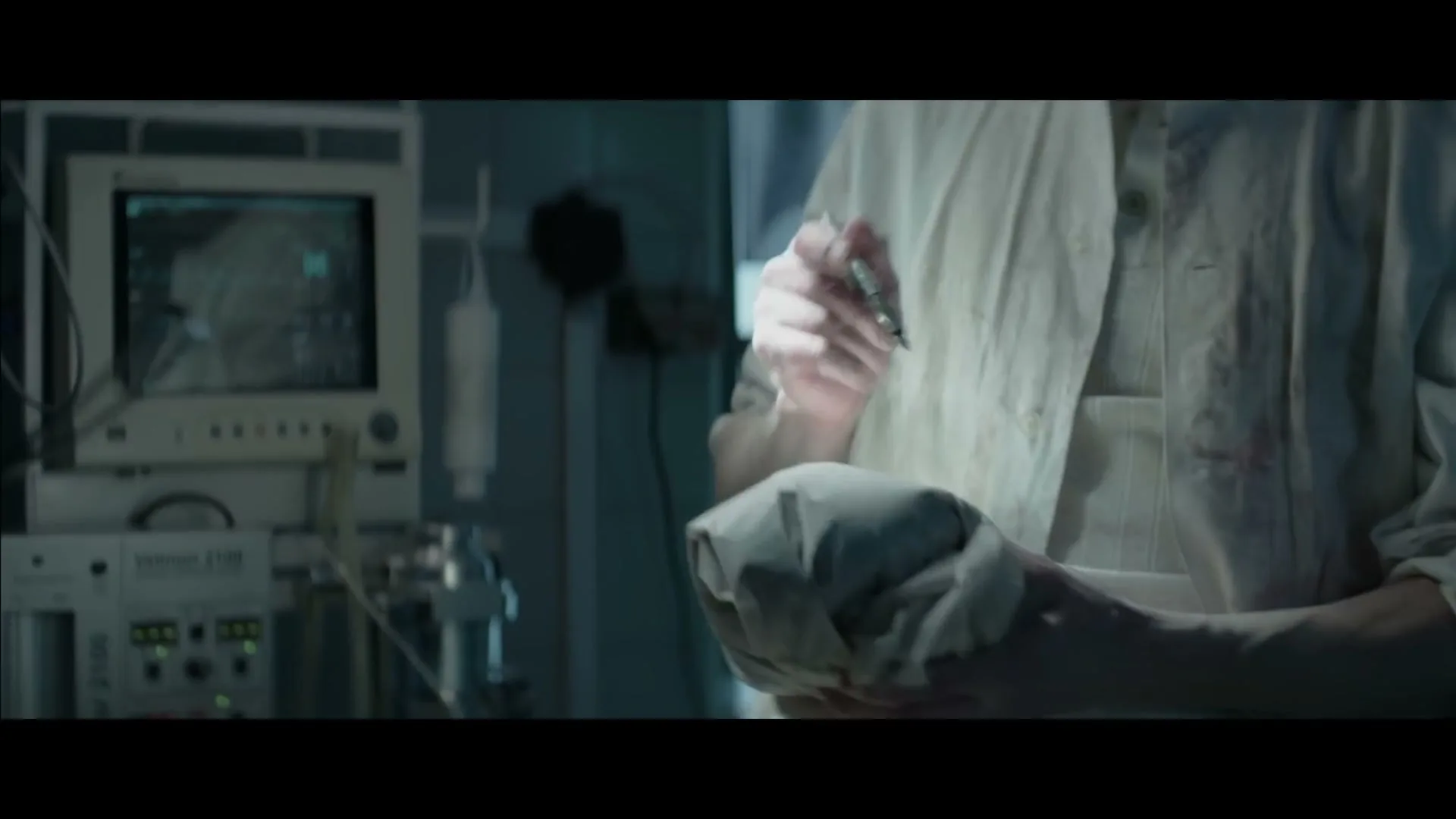 A person in a white lab coat with bloodstains on the sleeves is holding a small, wrapped object and a pen-like tool in a dimly lit room with medical equipment.