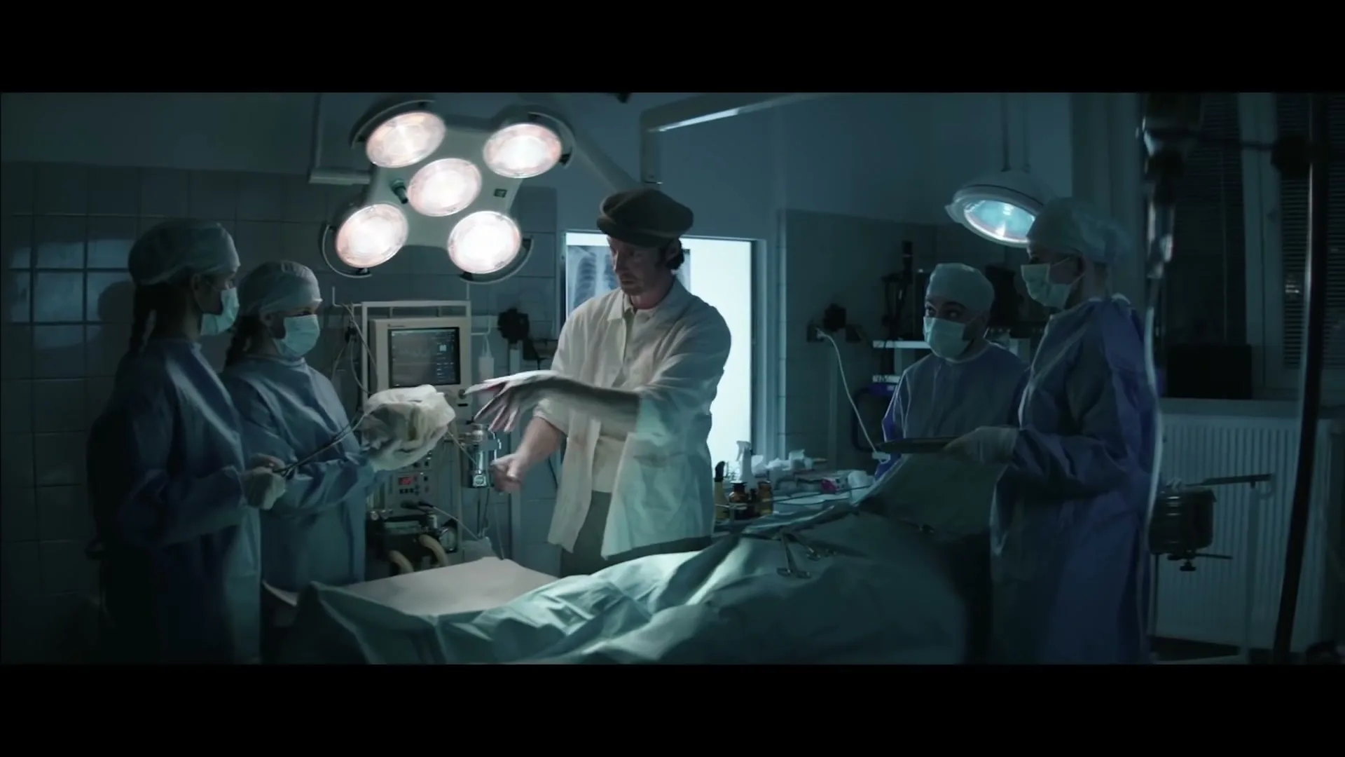 A man in a flat cap and white coat holds up a brain while four surgeons in scrubs and masks look on in a dimly lit operating room.