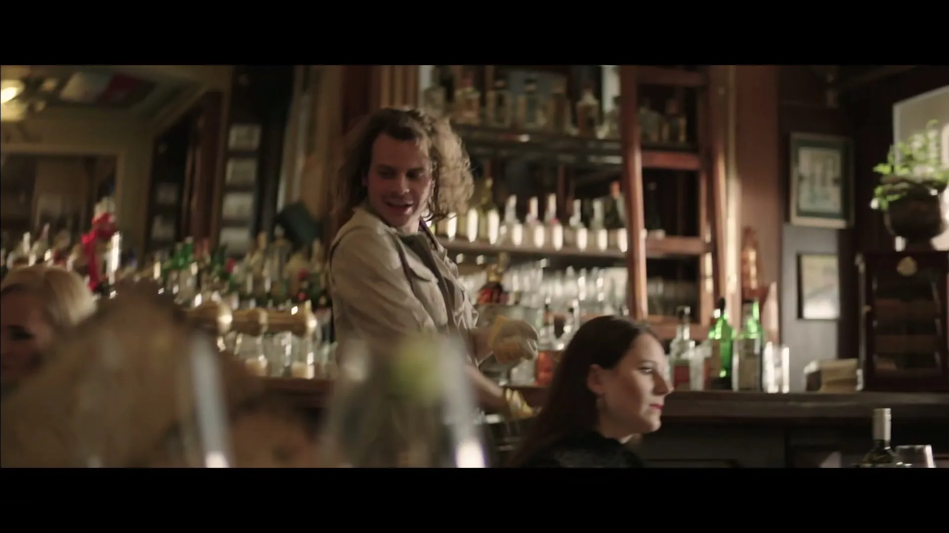 A waiter with curly hair and a beige jacket stands behind a bar, looking around and smiling, with bottles and glasses in the background.