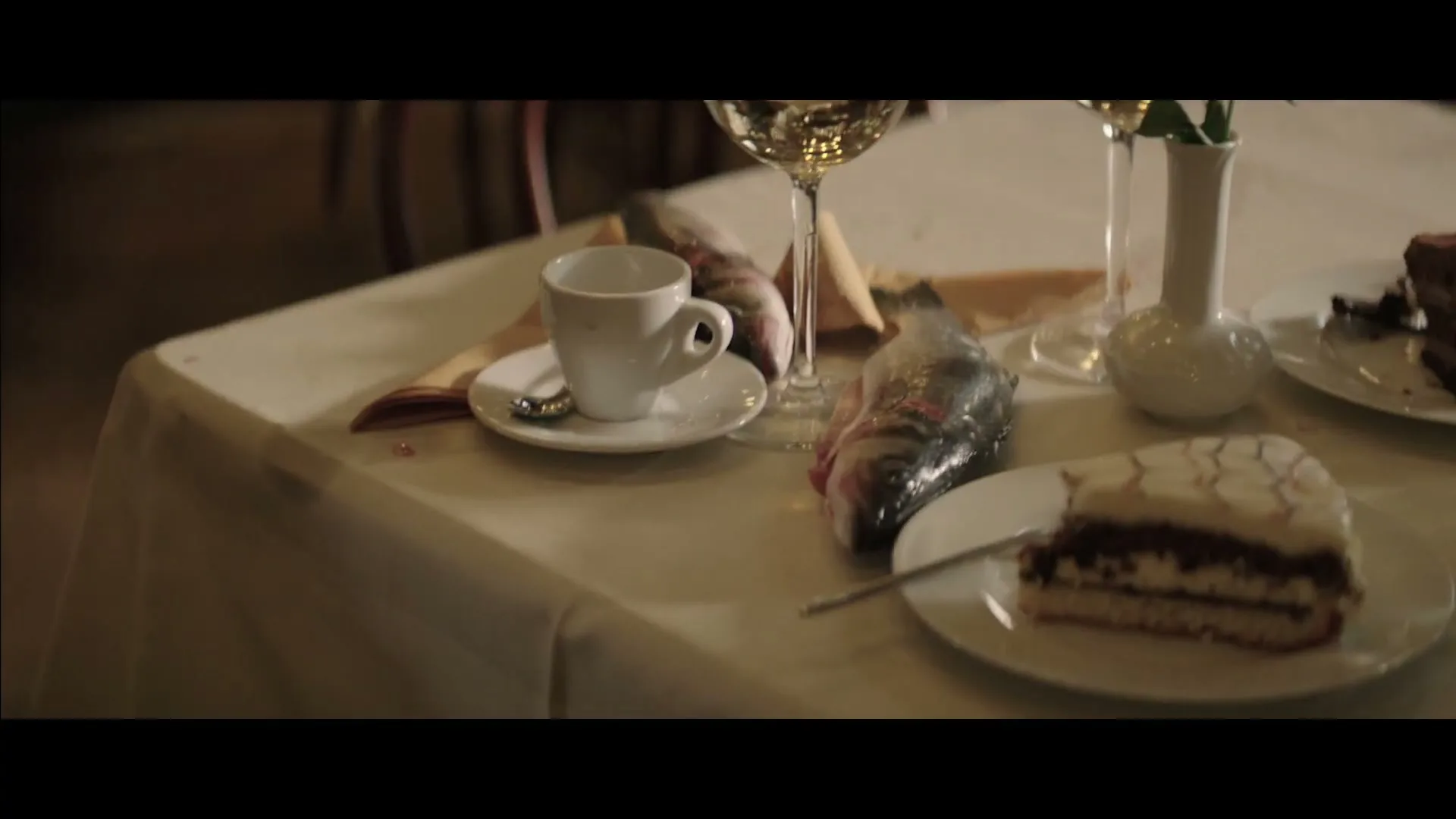 A close-up shot of a restaurant table with a white tablecloth, showing a coffee cup, wine glass, a whole fish, and a slice of cake.