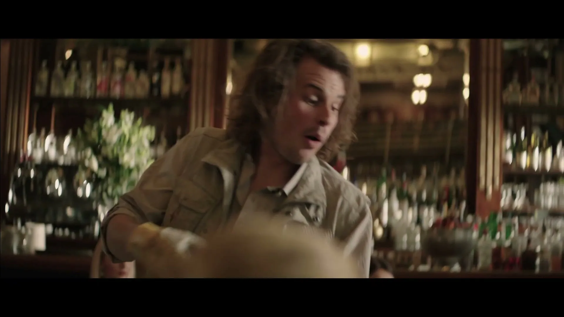 A young man with curly hair, wearing a beige jacket, looks surprised and leans forward over a table in a restaurant.