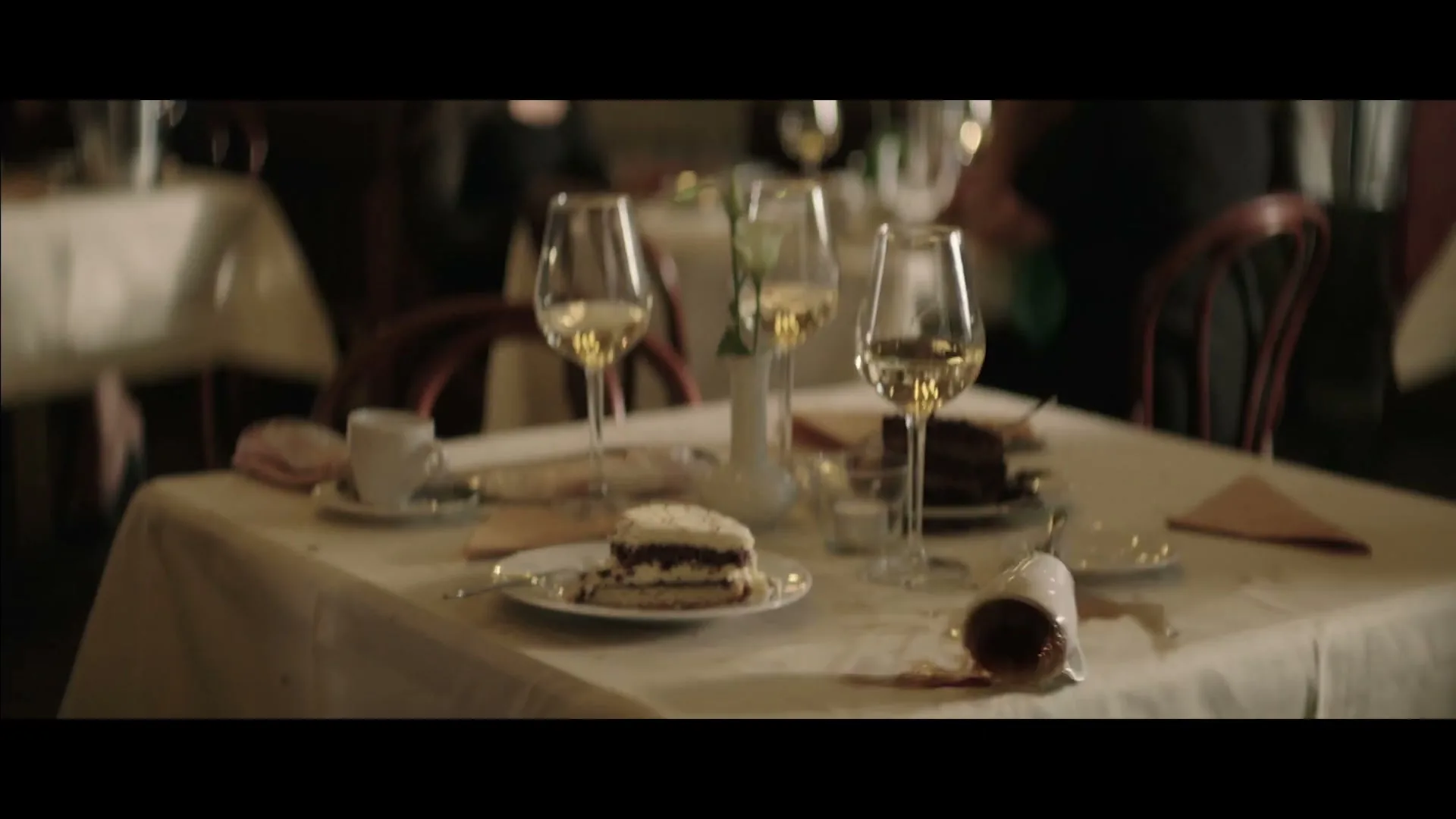 A coffee cup falls and spills on a white tablecloth next to a slice of cake and wine glasses in a restaurant.