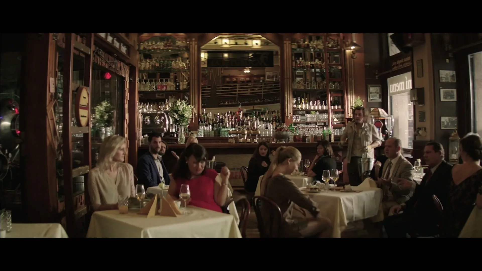 A waiter in a casual shirt stands behind a bar, while a woman in a red dress at a table in the foreground claps her hands, in a busy restaurant.