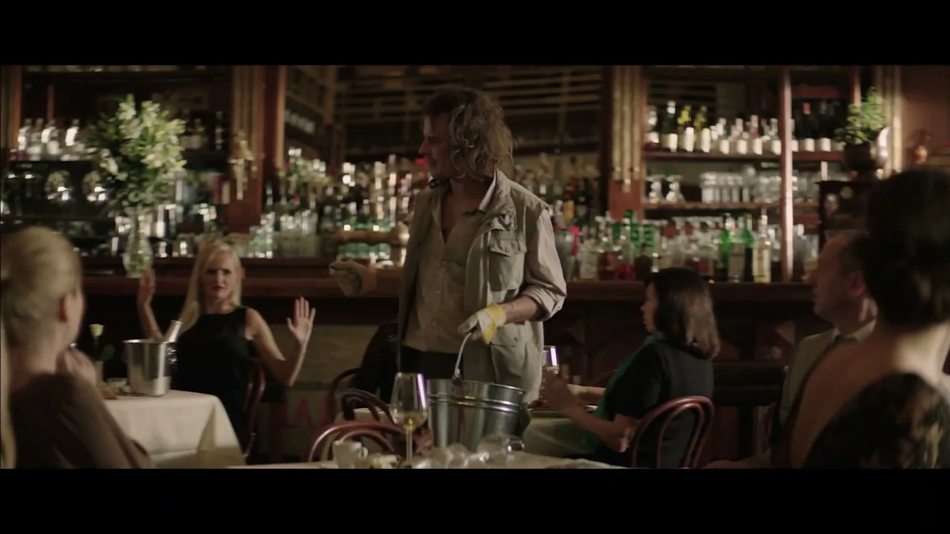 A male waiter with curly hair and a safari jacket pours water from a bucket into a glass at a fancy restaurant, as customers watch him.