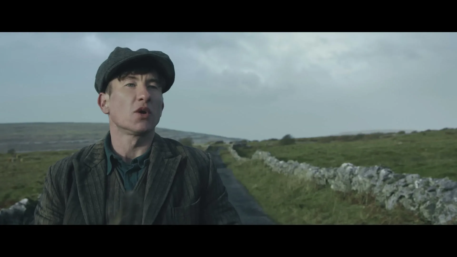 A man in a flat cap and tweed jacket walks along a narrow road with stone walls and green fields under a cloudy sky.