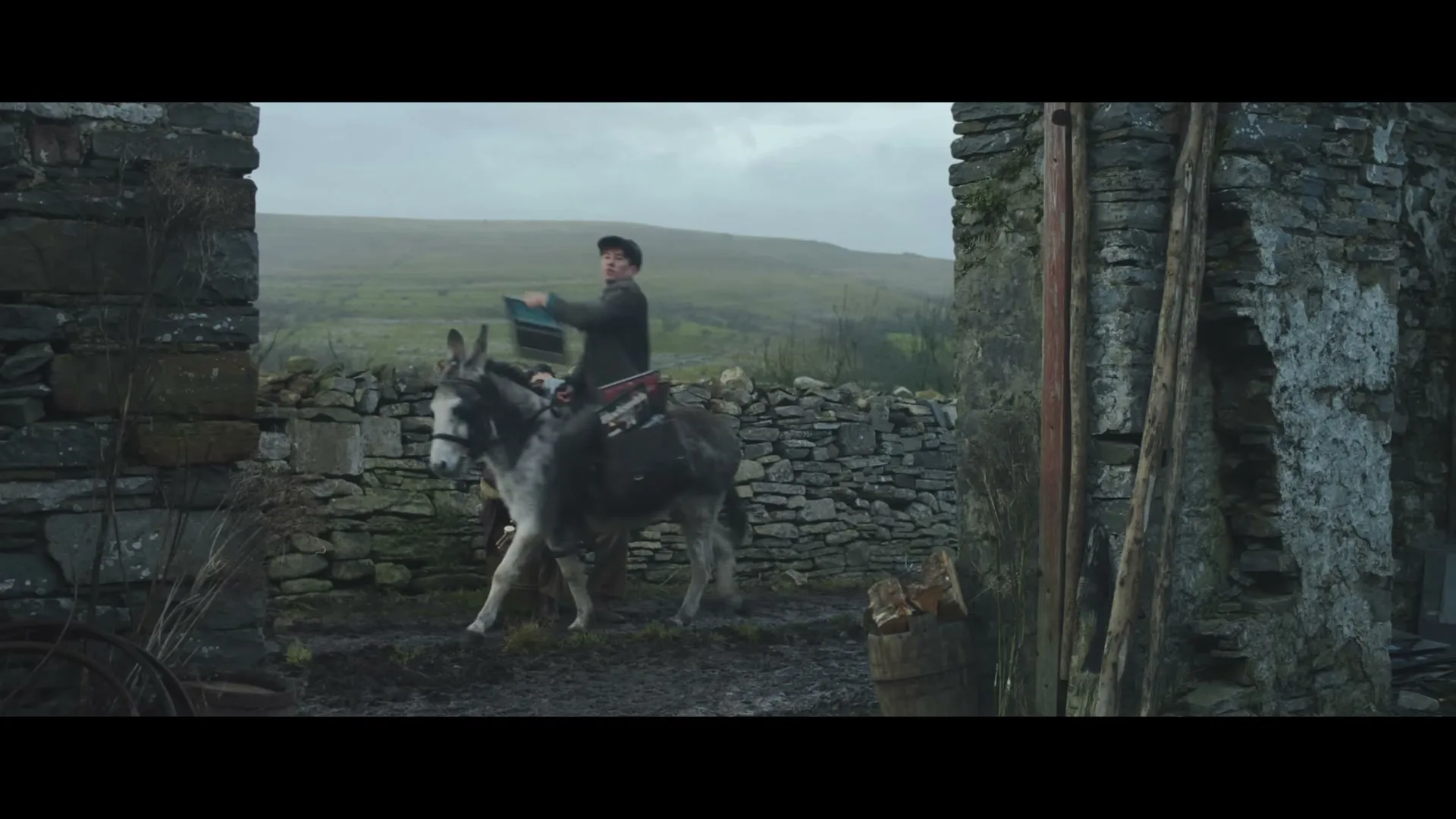 A young man in a cap rides a donkey through a stone archway, holding a book, in a rural, overcast landscape.
