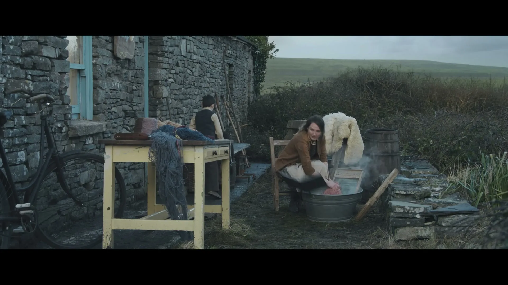 A young woman washes clothes in a tub outside a stone cottage, while a young man works nearby, in a rural setting.