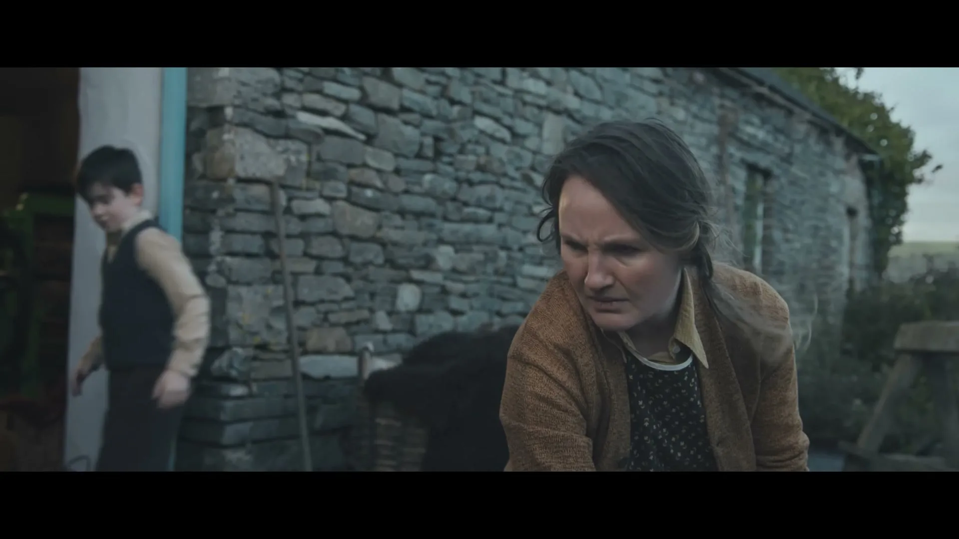 A woman with dark hair and a young boy are outside a stone building, the woman looks concerned while the boy walks away.
