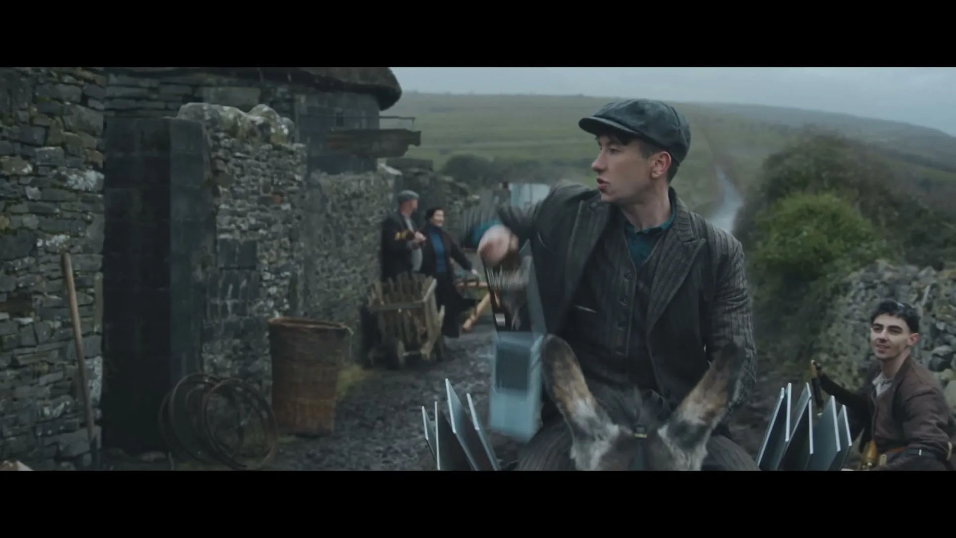 A young man in a flat cap rides a donkey, holding a laptop and a magazine, on a muddy path in a rural village.