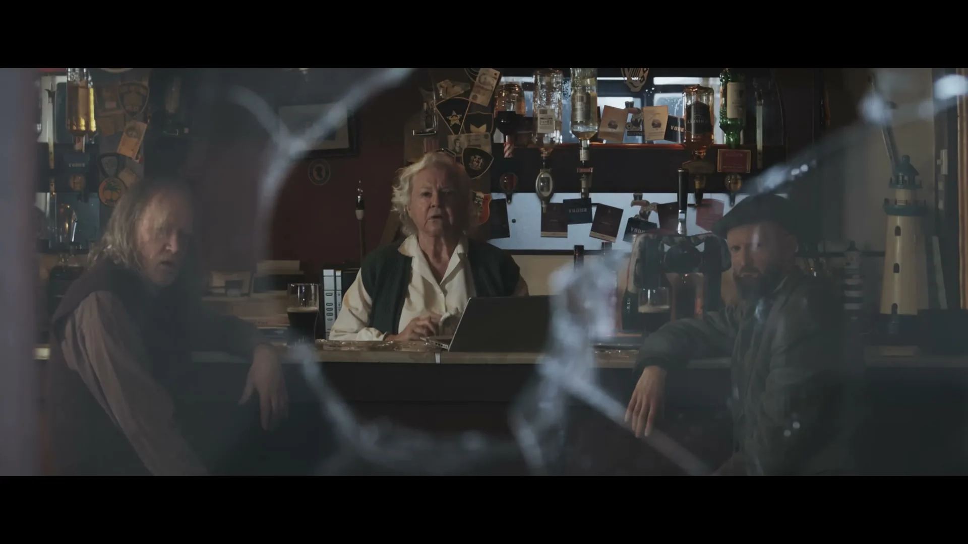 A woman with white hair sits behind a bar, looking at a laptop, flanked by two men, seen through a broken window in a dimly lit pub.
