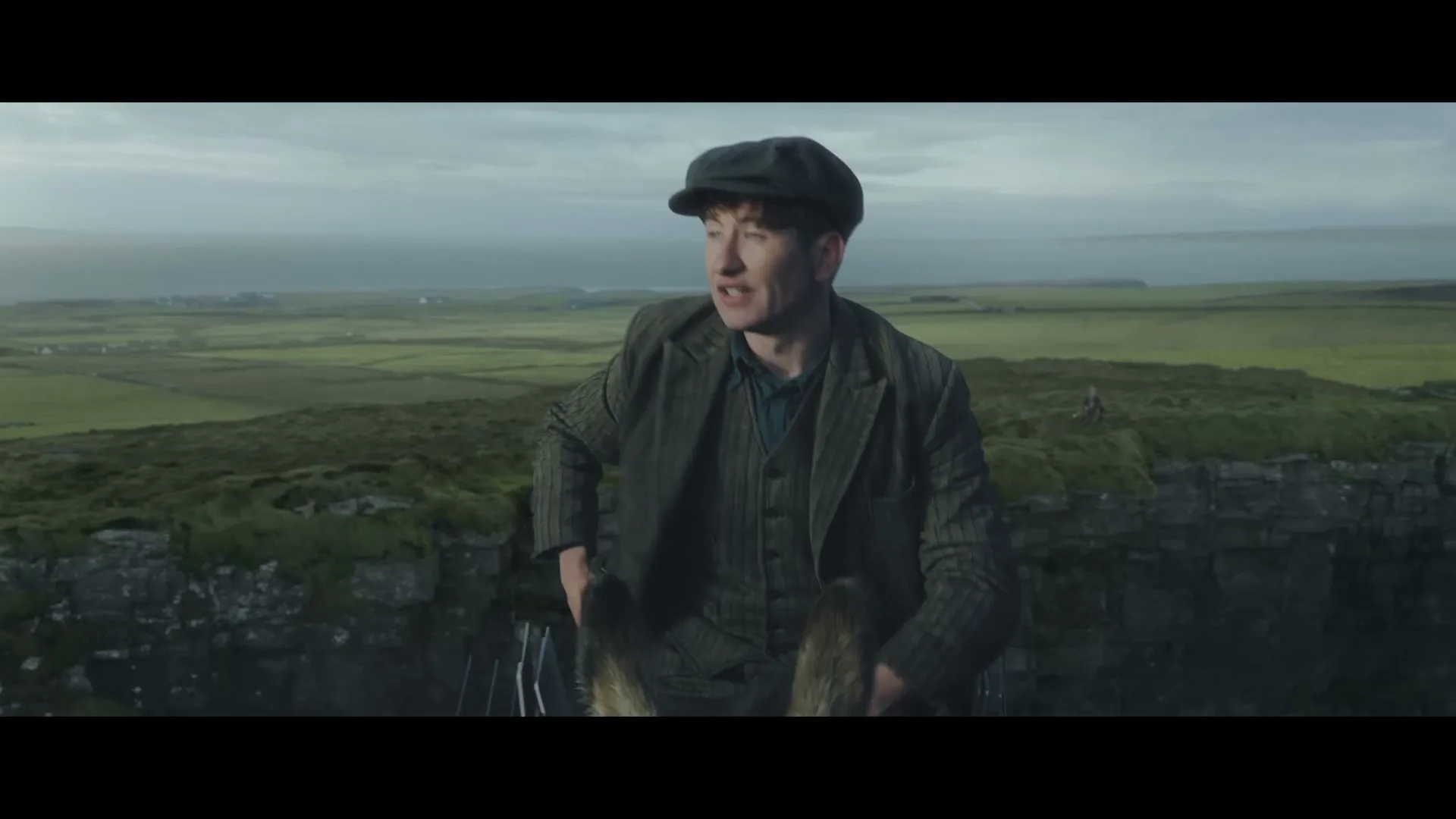 A young man in a tweed suit and flat cap sits on a cliff edge, looking around with a concerned expression, with green fields and a cloudy sky in the background.